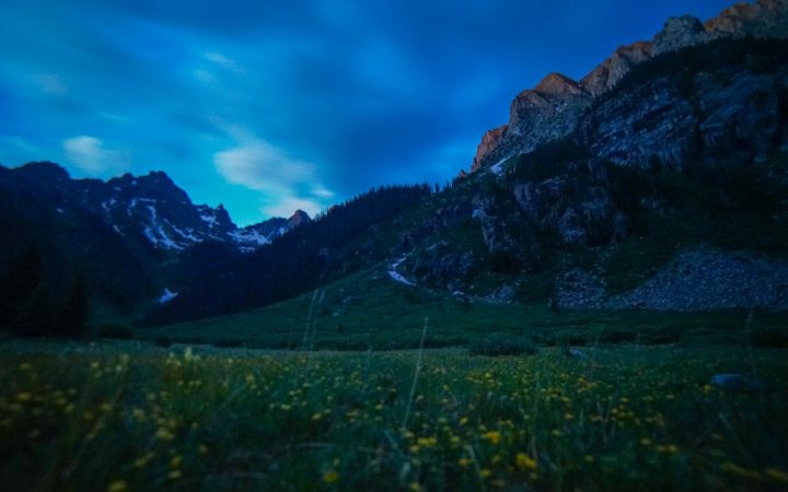 The sky is softly lit, illuminating a green meadow dotted with yellow wildflowers. There is a mountainous landscape surrounding the meadow.